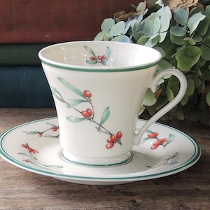 May include: A white teacup and saucer with a green rim and a floral design featuring red berries and green leaves.
