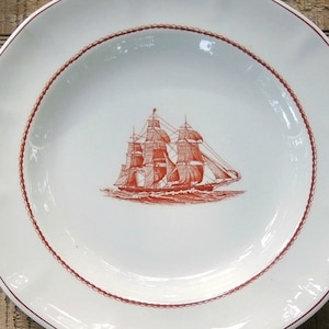 May include: A white oval ceramic plate with a red rope design border. The center of the plate features a red line drawing of a sailing ship.