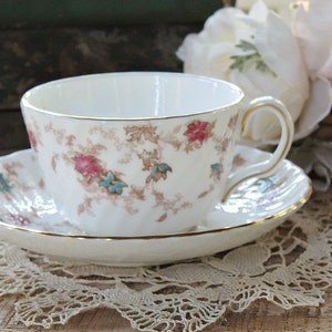 May include: A white teacup and saucer set with a floral pattern in pink, teal, and brown, accented with gold trim. The teacup sits on a matching saucer, resting on a lace doily. The set is a classic design.