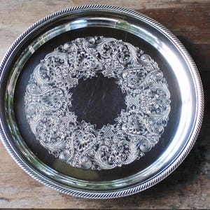 May include: A round, silver-colored serving tray with an ornate, embossed floral design in the center. The tray has a rope-like border and is set against a wooden surface.