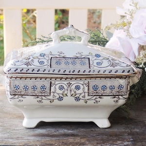 May include: A white porcelain serving dish with a lid. The dish has a blue and gold floral pattern. The dish is on a wooden surface.