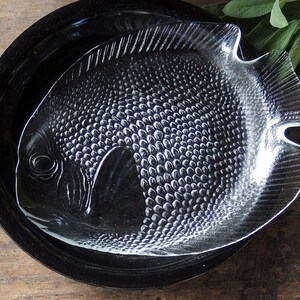 May include: A clear glass serving dish shaped like a fish with a textured surface. The dish is sitting on a black plate.