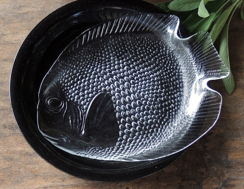 May include: A clear glass serving dish shaped like a fish with a textured surface. The dish is sitting on a black plate.
