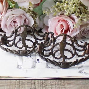 May include: Two antique bronze drawer pulls with ornate scrollwork, resting on sheet music with a floral arrangement of pink roses and greenery in the background. The pulls have a curved design with a central handle.