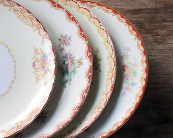 May include: Four antique porcelain plates with floral and gold trim. The plates have a white center with a delicate floral pattern and a scalloped edge with red and gold accents. The plates are arranged on a wooden surface.