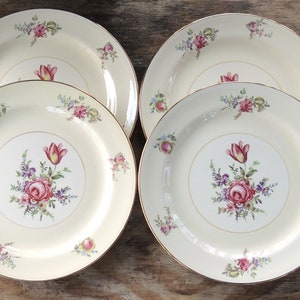 May include: Set of four white ceramic dinner plates with a gold rim and floral design. The plates feature a pink rose, purple and white flowers, and green leaves.