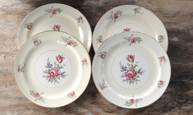 May include: Set of four white ceramic dinner plates with a gold rim and floral design. The plates feature a pink rose, purple and white flowers, and green leaves.