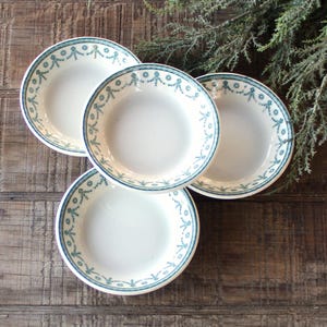 May include: Four white ceramic plates with a teal floral design around the rim. The plates are arranged on a rustic wooden surface, with a sprig of greenery in the background. The plates are a classic design.