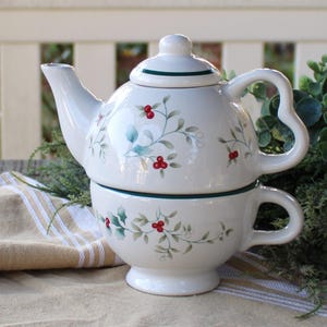 May include: A white ceramic teapot and teacup set with a green trim and a holly berry design. The teapot has a lid and the teacup has a handle.