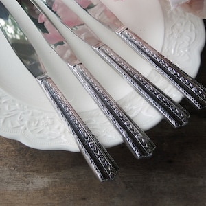 May include: Four silver knives with ornate handles rest on a white, patterned plate. The knives have a shiny, reflective blade and a detailed, textured handle design. The plate is white with a floral pattern.