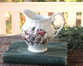 以下が含まれることがあります： ピンク、茶色、青色の花柄が施された白いセラミックピッチャー。ピッチャーには曲がったハンドルと注ぎ口があります。ヴィンテージの緑色の本の上に置かれています。