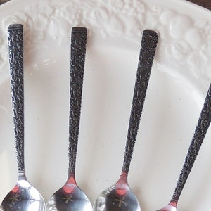 May include: Four silver spoons with ornate, dark gray floral patterns on the handles rest on a white, embossed plate. The spoons have a classic design, with a small star-shaped detail on the bowl.