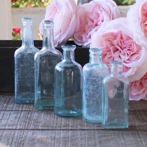May include: Five antique, clear glass bottles of varying shapes and sizes are arranged on a weathered wooden surface. Soft pink roses are in the background. The bottles have a slight blue-green tint.