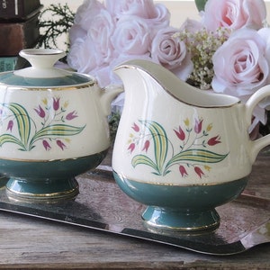 May include: A vintage cream-colored sugar and creamer set with a green base and gold trim. The set features a floral design with pink and yellow flowers. The set is on a silver tray.