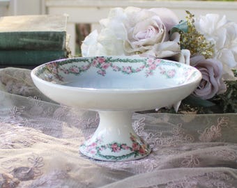 May include: A white ceramic pedestal bowl with a floral design of pink roses and green leaves. The bowl has a wide, shallow basin and a short, rounded pedestal base. The floral pattern encircles the rim and base of the bowl.