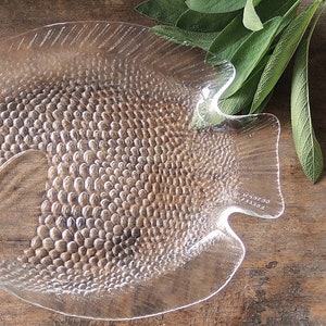 May include: A clear glass fish-shaped serving dish with a textured surface. The dish has a raised rim and a flat bottom. The dish is made of glass and has a clear finish.