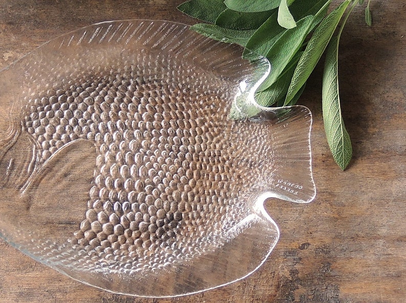 May include: A clear glass fish-shaped serving dish with a textured surface. The dish has a raised rim and a flat bottom. The dish is made of glass and has a clear finish.