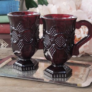 May include: Two ruby red glass mugs with a textured design and a handle. The mugs are on a silver tray.