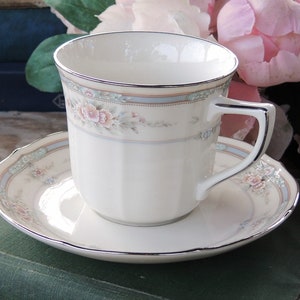 May include: A white porcelain teacup and saucer with a floral pattern in shades of pink, blue, and green. The cup and saucer have a silver trim.