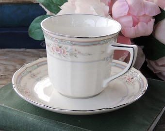 May include: A white porcelain teacup and saucer with a floral pattern in shades of pink, blue, and green. The cup and saucer have a silver trim.