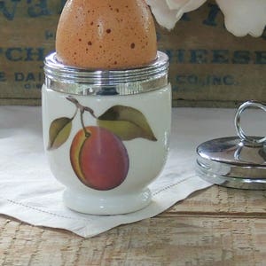 May include: A white ceramic egg cup with a silver lid and a painted plum design. The egg cup is filled with a brown egg. A silver egg cup lid with a ring handle is also shown.
