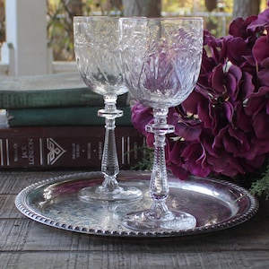 May include: Two clear crystal wine glasses with intricate etched designs sit on a silver serving tray. The glasses have detailed stems and bowls. A bouquet of deep purple flowers and a stack of vintage books are in the background.