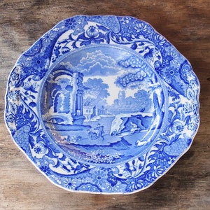May include: A blue and white porcelain plate with a decorative floral border and a central scene of a landscape with a bridge and trees.