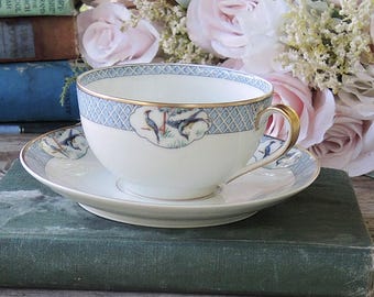 May include: A white teacup and saucer with a blue and gold floral design. The teacup has a gold rim and a handle. The saucer has a matching design. The teacup and saucer are sitting on a stack of books.