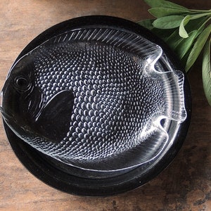 May include: A clear glass serving dish shaped like a fish with a textured surface. The dish is sitting on a black circular base.