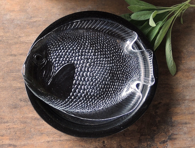 May include: A clear glass serving dish shaped like a fish with a textured surface. The dish is sitting on a black circular base.