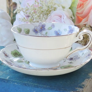 May include: A white teacup and saucer with a floral pattern of purple flowers and green leaves. The teacup and saucer are sitting on a blue book.