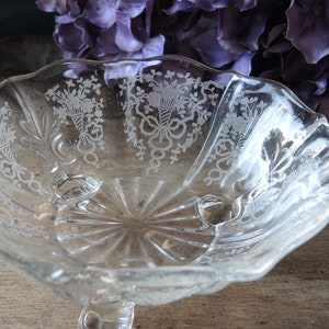 May include: Clear glass footed bowl with etched floral and ribbon designs. The bowl has a scalloped edge and a starburst pattern on the base. A decorative piece for serving or display.