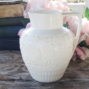 May include: White ceramic pitcher with an embossed floral design. The pitcher has a wide mouth and a handle.