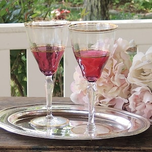 May include: Two stemmed wine glasses filled with a red liquid, set on a silver tray. The glasses have clear stems and bowls with gold rims. The tray is oval with a decorative border. Pink and white flowers are in the background.