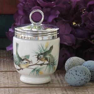 May include: A white porcelain egg cup with a silver lid and handle, decorated with a detailed illustration of two birds on a branch. Three speckled blue eggs are next to the egg cup. The background includes purple flowers and a wooden surface.