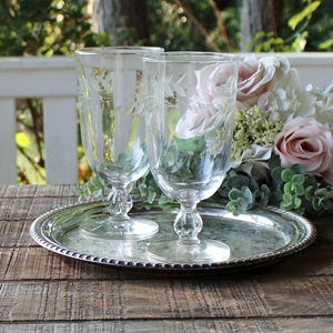 May include: Two clear, etched glass goblets with floral designs sit on a silver-colored, beaded-edge tray. The goblets have a rounded base and a long stem. A bouquet of pink and white flowers is in the background.