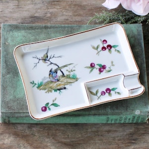 May include: A rectangular white porcelain ashtray with a gold rim, featuring a painted scene of birds on a branch and sprigs of berries. The ashtray has a small rectangular section and a curved area for resting cigarettes.