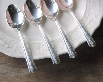 May include: Four silver spoons with ornate handles are arranged on a white plate with a decorative pattern. The spoons are polished and reflect light.