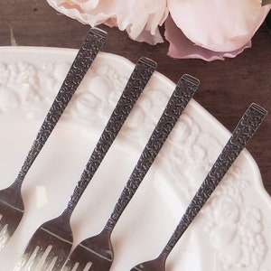 May include: Four silver forks with an ornate design on a white plate with a floral pattern.