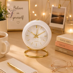 Può includere: Un orologio analogico rotondo bianco con una base circolare dorata e lancette dorate. Il quadrante presenta indicatori dorati per le ore e la scritta QUARTZ stampata in nero. L'orologio ha una finitura liscia e lucida e un design sferico.