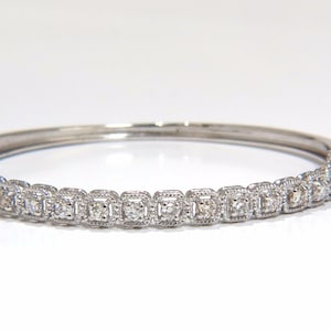 Könnte beinhalten: Ein silberfarbenes Klapparmband mit einer durchgehenden Reihe quadratischer Fassungen, die jeweils einen kleinen runden, klaren Stein enthalten. Der Metallrahmen weist an den Kanten jedes quadratischen Glieds eine feine Perlenstruktur auf.