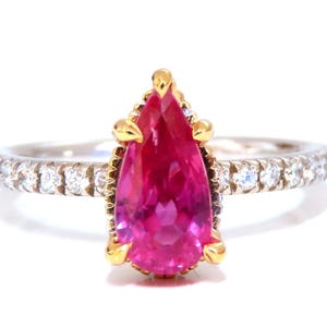 Puede incluir: Un anillo con una piedra preciosa de color rosa vibrante en forma de pera, engastada en garras de oro amarillo. La banda es de metal plateado y está adornada con una serie de pequeñas piedras redondas y transparentes incrustadas a ambos lados.