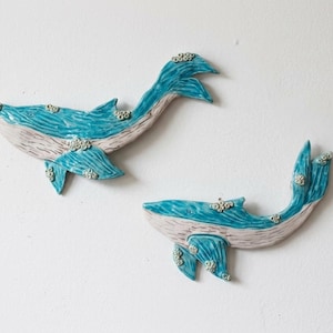 Op de afbeelding: Twee keramische wandsculpturen in de vorm van walvissen. Elke walvis heeft een fel turquoise blauw bovenlichaam met textuurdetails en een lichtbeige onderbuik met lineaire markeringen. Kleine witte cirkelvormige accenten zitten op de vinnen.