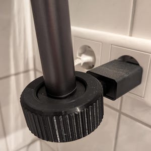 Peut inclure: Un appareil cylindrique noir avec une base circulaire texturée et striée est branché dans une prise murale blanche. Une tige noire verticale s'étend vers le haut depuis le centre de la base circulaire, reliée à l'adaptateur secteur mural.