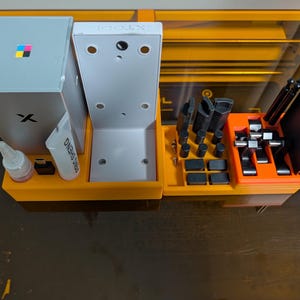 Op de afbeelding: Een oranje kunststof organizerbak met diverse accessoires voor lasergraveermachines, waaronder een zilverkleurige doos met logo, een witte verticale beugel met het opschrift xTool, een kleine tube, een USB-stick en diverse zwarte metalen onderdelen.