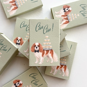 May include: A collection of rectangular light green cards featuring an illustration of a brown and white Cavalier King Charles Spaniel wearing a black bow tie. The dog carries a stacked tower of champagne glasses. The text Cin Cin! is printed.