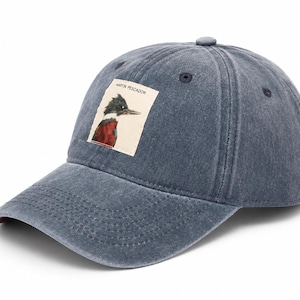 Puede incluir: Una gorra de béisbol de color azul marino desgastado con un parche rectangular en la parte delantera. El parche muestra la ilustración de un pájaro con cresta oscura, pico largo y pecho rojizo, con el texto MARTIN PESCADOR impreso encima.