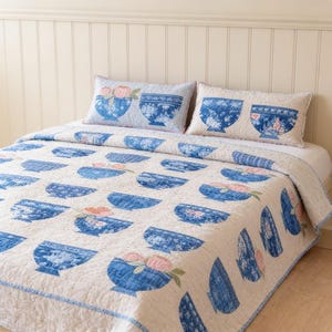 May include: A cream-coloured quilt and two matching rectangular pillows featuring a repeating pattern of blue and white bowls filled with pink flowers. The bedding set has a quilted texture and a consistent print of floral-filled vessels throughout.