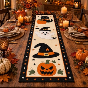 Puede incluir: Un camino de mesa rectangular con un centro de color crema que presenta cuatro ilustraciones de Halloween: un gato negro con una calabaza, un fantasma blanco con sombrero de bruja, un gran sombrero de bruja negro y una calabaza tallada.