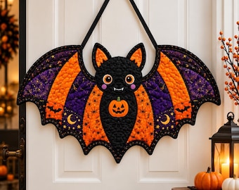 Könnte beinhalten: Ein gesteppter Wandbehang in Fledermausform aus schwarzem, orangefarbenem und violettem Stoff. Die Fledermaus hat ein zentrales Kürbis-Motiv, spitze Ohren und ein schwarzes Band zum Aufhängen. Die Flügel sind mit Sternen und Monden verziert.